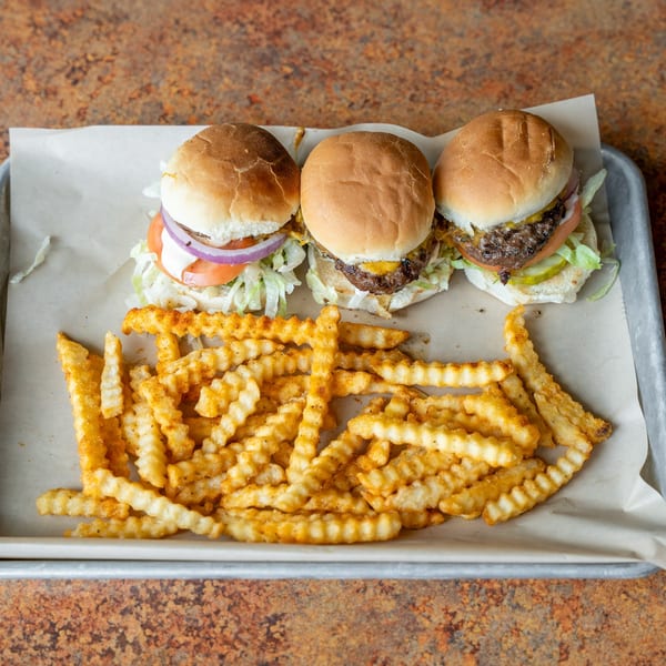 Three sliders with cheese, lettuce, tomato, onion, and pickles, served with crinkle-cut fries on a tray.
