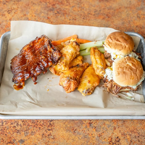 A tray of BBQ ribs, chicken wings, and pulled pork sliders with celery and carrots.